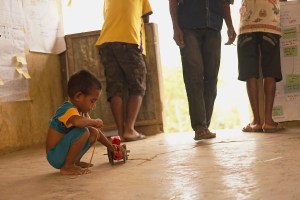 A child plays off to the side of the community Action Planning with his home made toy car. Community Action Planning happens in the community. Life in the community does not stop. For two days the meetings become a part of the community. Often while parents and grandparents discuss nutrition issues and solutions children are playing off to the side or even joining in the discussion. The charts in the background are a nutritional calendar, sunshine wheel (reflecting nutritional themes), and a table that people discuss how women and men can take different roles in helping children and new mothers increase their nutrition. 